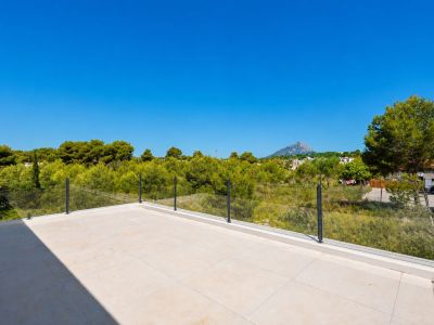 Amplia terraza superior con suelo de gres porcelánico claro y barandilla de cristal, ofreciendo vistas a la vegetación mediterránea y al perfil de la montaña.