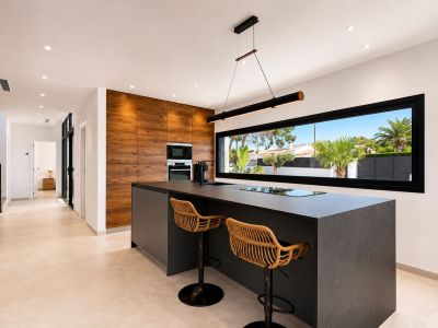 Cocina moderna con isla central de encimera oscura y taburetes de ratán. Pared de madera con electrodomésticos integrados y ventanal horizontal con vistas al exterior.