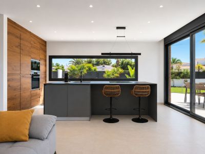 Cocina moderna con isla central, equipada con electrodomésticos integrados y barra con taburetes de ratán. Grandes ventanales ofrecen vistas al jardín y acceso directo al exterior.
