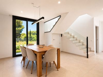 Comedor moderno con mesa de madera clara y seis sillas tapizadas, bajo una lámpara colgante lineal. Junto a él, una escalera contemporánea con peldaños iluminados y barandilla de cristal.