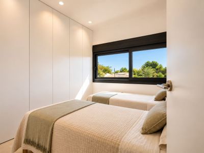 Dormitorio secundario con dos camas individuales, armarios empotrados blancos sin tiradores y ventana con vistas a la vegetación mediterránea. Iluminación empotrada y luz natural.