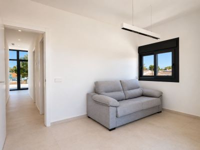 Espacio auxiliar luminoso con sofá gris, ventana de marco negro y vistas al entorno. El pasillo adyacente se abre a una gran ventana exterior, destacando la luz natural.