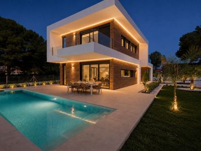 Exterior de la villa moderna al anochecer, con líneas arquitectónicas definidas, revestimiento de efecto madera y cálida iluminación LED perimetral. Piscina privada y terraza comedor exterior iluminadas, rodeadas de jardín.
