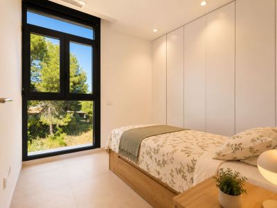 Luminoso dormitorio con cama individual de base de madera, armarios empotrados blancos y gran ventanal negro con vistas a la naturaleza. Iluminación empotrada en el techo.