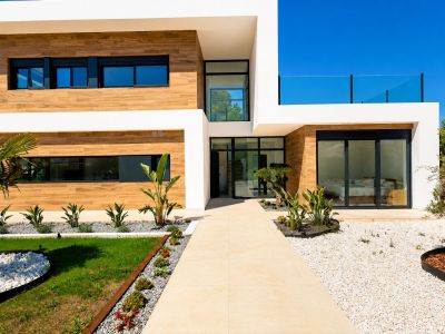 Moderna fachada de villa con revestimiento efecto madera, paredes blancas y camino embaldosado hacia la entrada principal acristalada, con paisajismo mediterráneo minimalista.