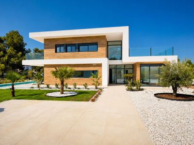 Vista exterior de la villa moderna con fachada de dos plantas, revestimiento efecto madera y grandes ventanales. Caminos de baldosas, jardín con palmeras y olivo, y piscina visible.