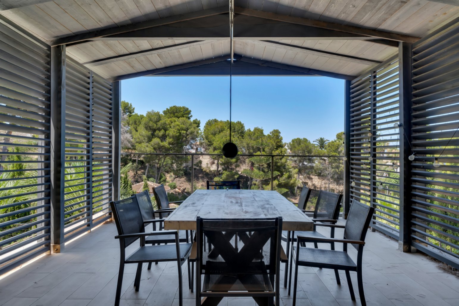 Amplia terraza cubierta con comedor exterior, mesa de madera rústica y sillas oscuras, enmarcada por celosías que ofrecen privacidad y vistas despejadas a la vegetación mediterránea.
