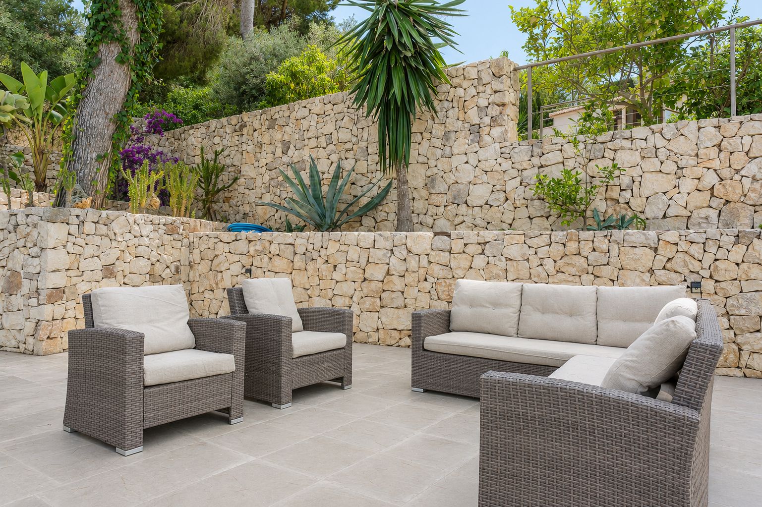 Amplia terraza exterior con mobiliario de ratán y cojines claros, enmarcada por muros de piedra seca escalonados y exuberante vegetación mediterránea.