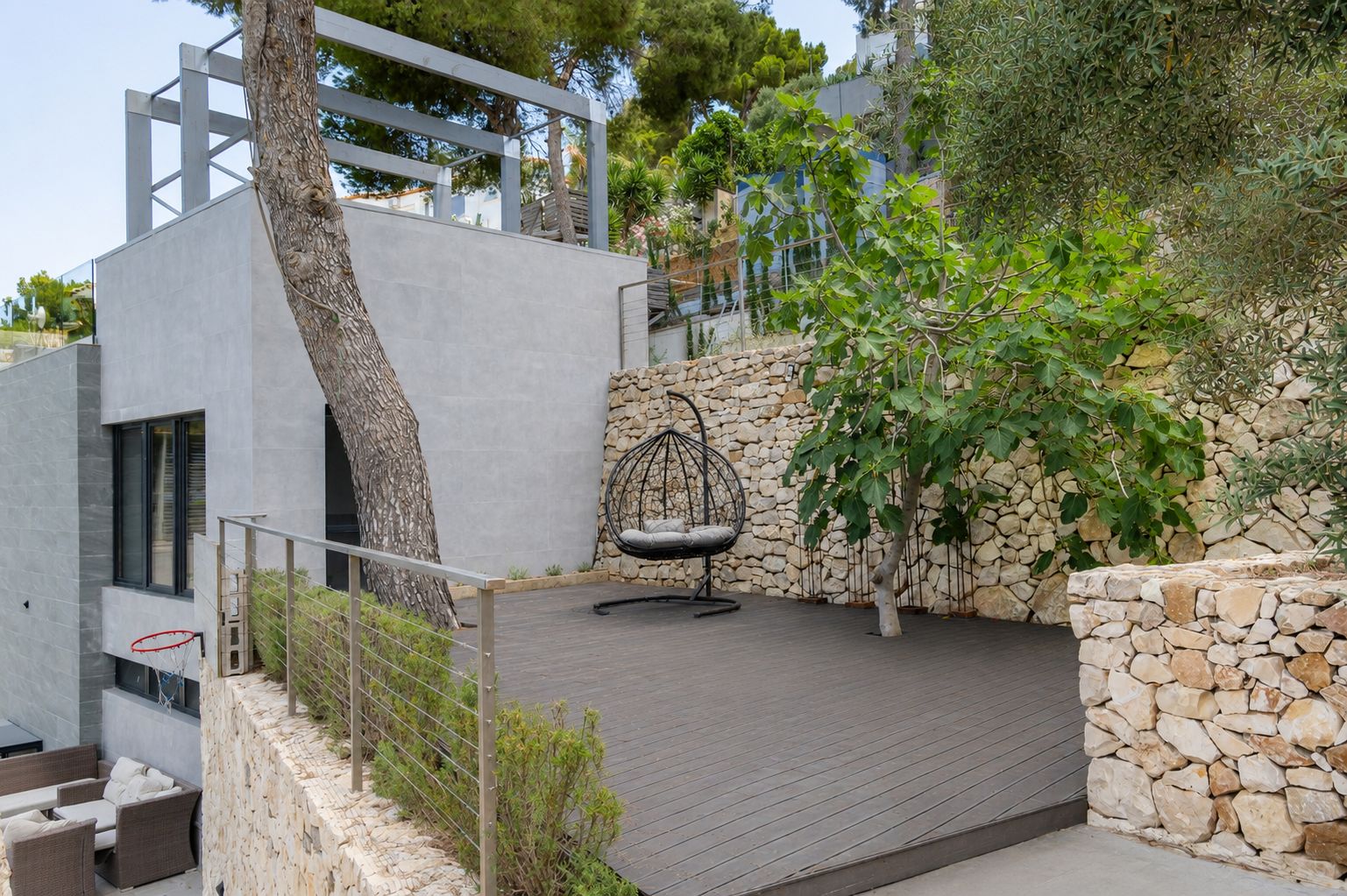 Amplia terraza exterior con tarima de composite oscuro, equipada con un sillón colgante y rodeada de muros de piedra natural. Árboles mediterráneos y arbustos aportan privacidad y se integran con la arquitectura moderna.