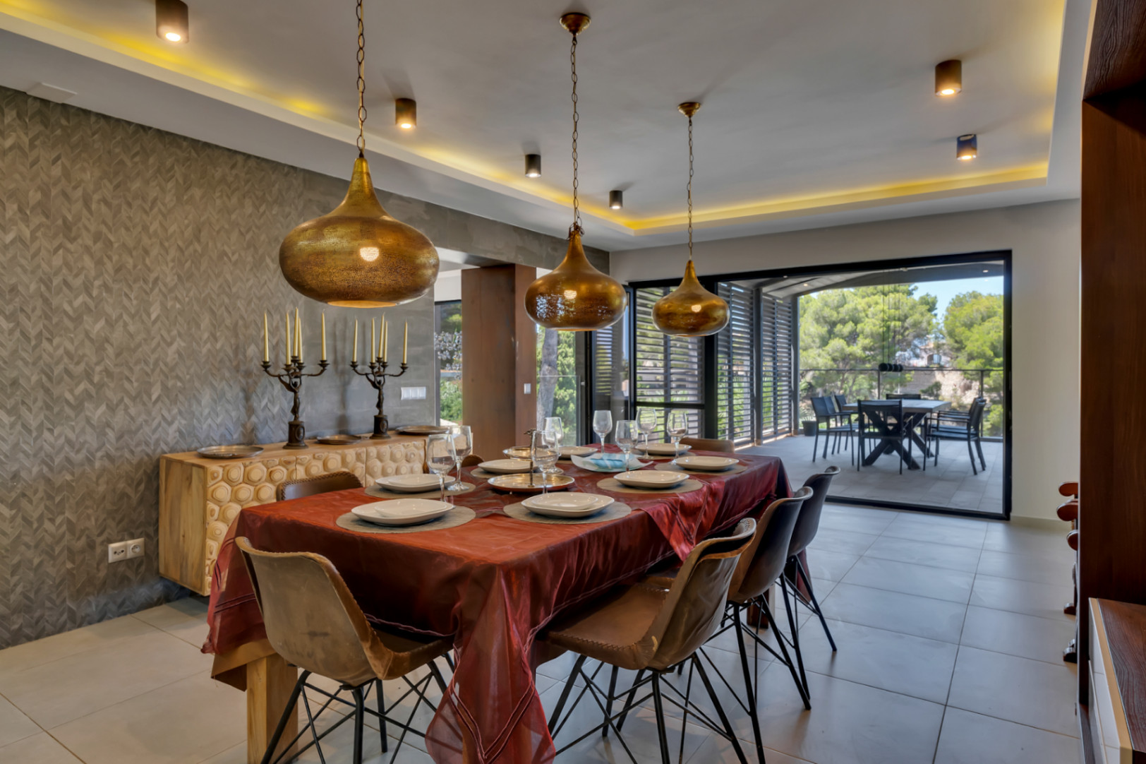 Amplio comedor con mesa de madera y sillas modernas, iluminado por tres lámparas colgantes doradas y luz indirecta. La pared texturizada y el aparador tallado añaden carácter, con acceso a una terraza exterior.