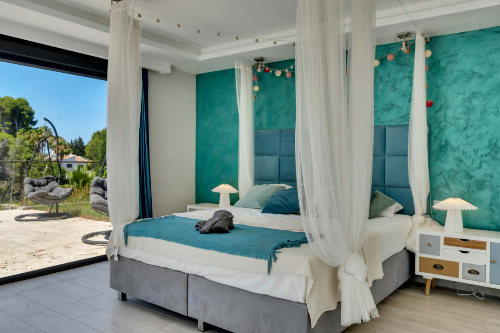 Amplio dormitorio con cama con dosel y cabecero tapizado azul, pared decorativa turquesa texturizada y acceso directo a terraza exterior con sillas colgantes y vegetación.