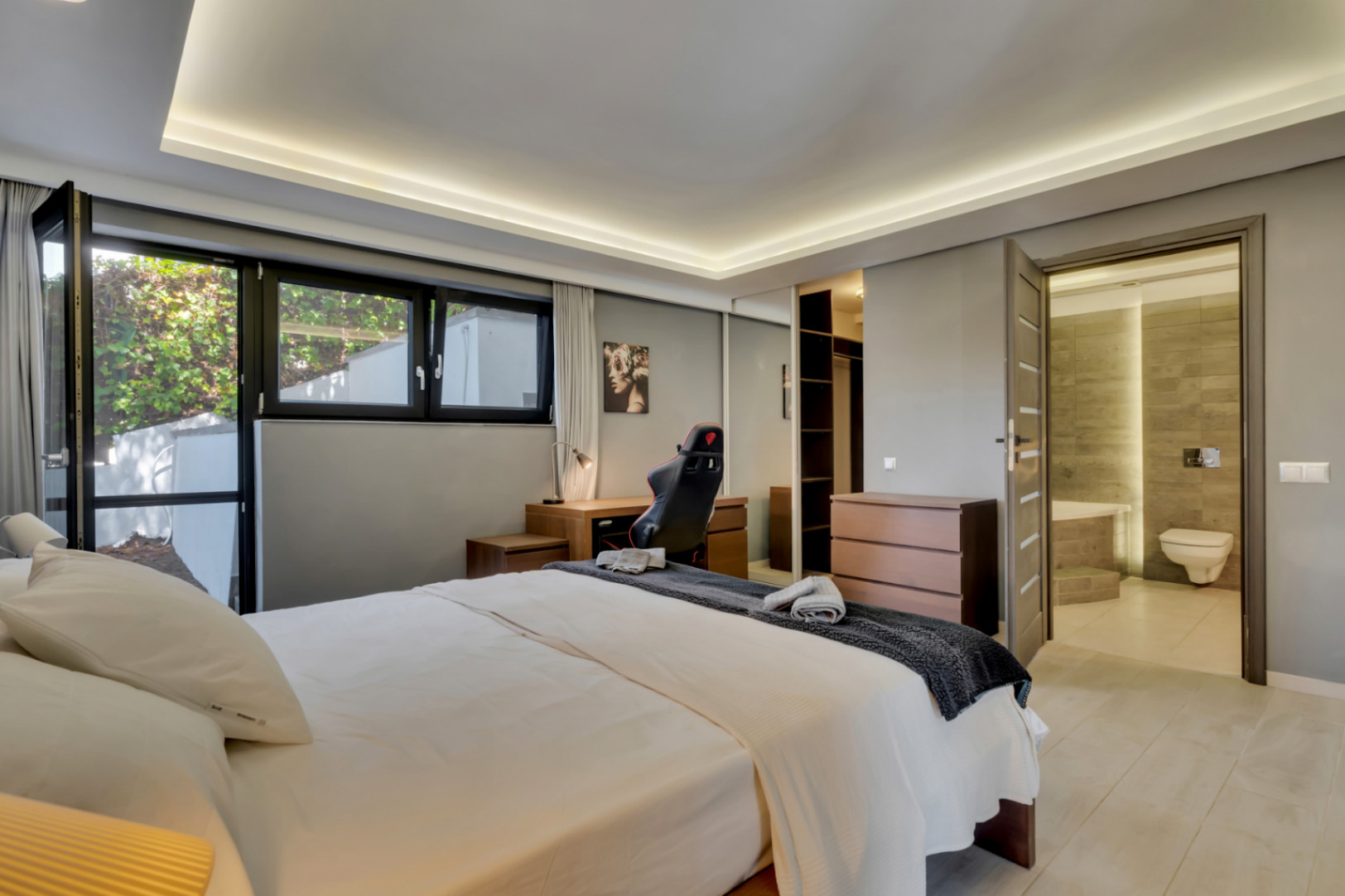Amplio dormitorio con cama doble, escritorio de madera y ventanales que dan al exterior. Incluye armario con espejo, cómoda y acceso directo a un baño en suite con azulejos contemporáneos.