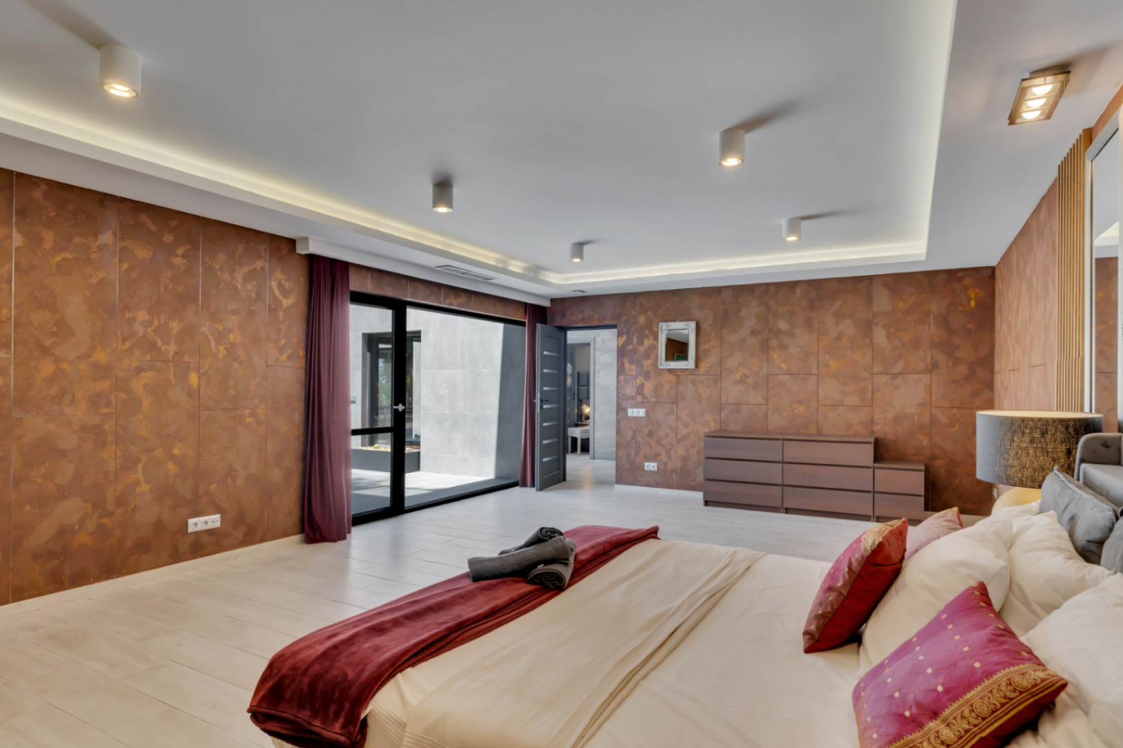 Amplio dormitorio con paredes de acabado cobrizo texturizado y suelo claro. Dispone de acceso directo a un patio exterior a través de una gran puerta corredera de cristal y una iluminación de techo moderna.