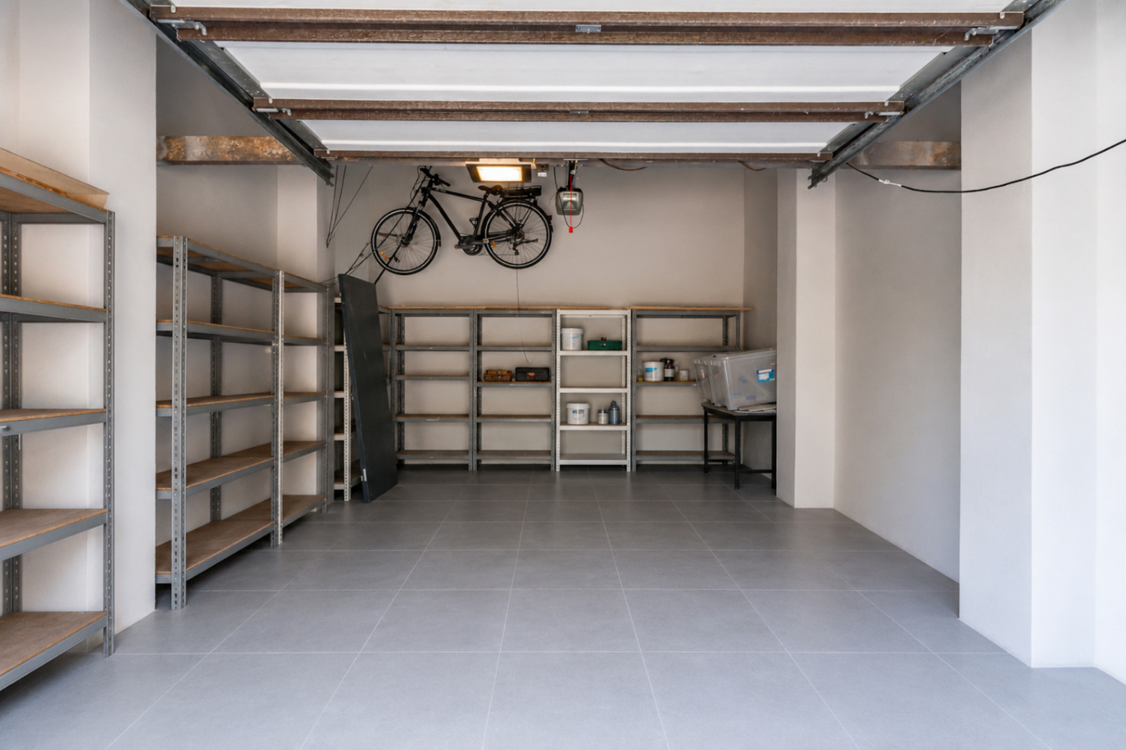 Amplio garaje con suelo de baldosas grises, extensas estanterías metálicas para almacenamiento organizado y soporte para bicicleta en pared. La estructura del techo expuesta ofrece un toque funcional.