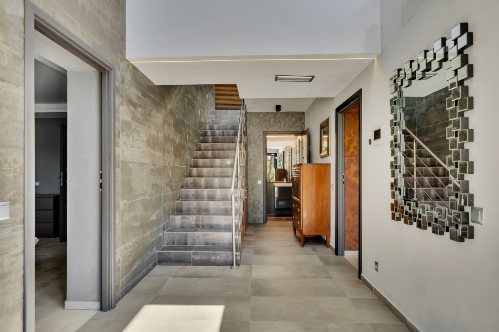 Amplio recibidor con escalera de diseño, revestida con azulejos de efecto hormigón y barandilla de acero inoxidable. El suelo es de gres porcelánico gris y un espejo modular decora la pared.