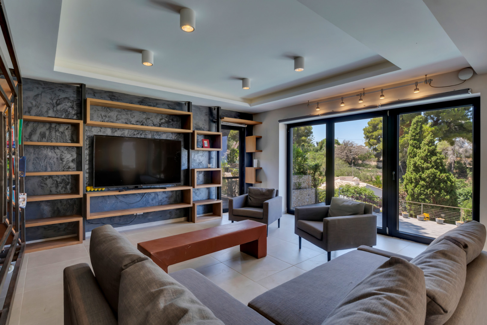 Amplio salón con estanterías de madera a medida y TV integrada en pared texturizada. Grandes ventanales ofrecen vistas verdes, junto a cómodos sofás grises y suelo de baldosas claras.