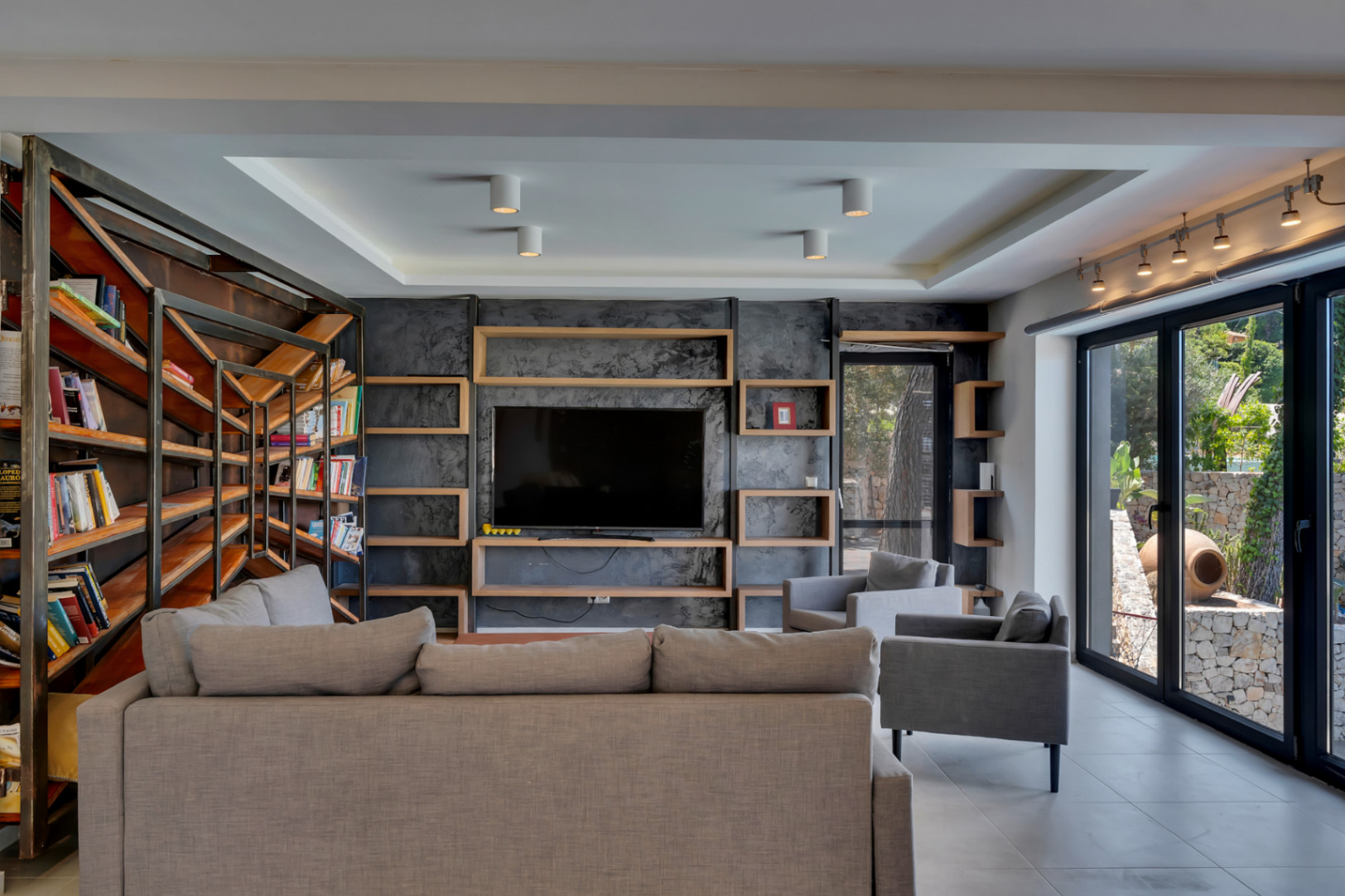Amplio salón con pared de TV en acabado texturizado oscuro y estantes de madera, complementado por una estantería industrial y ventanales al jardín mediterráneo.