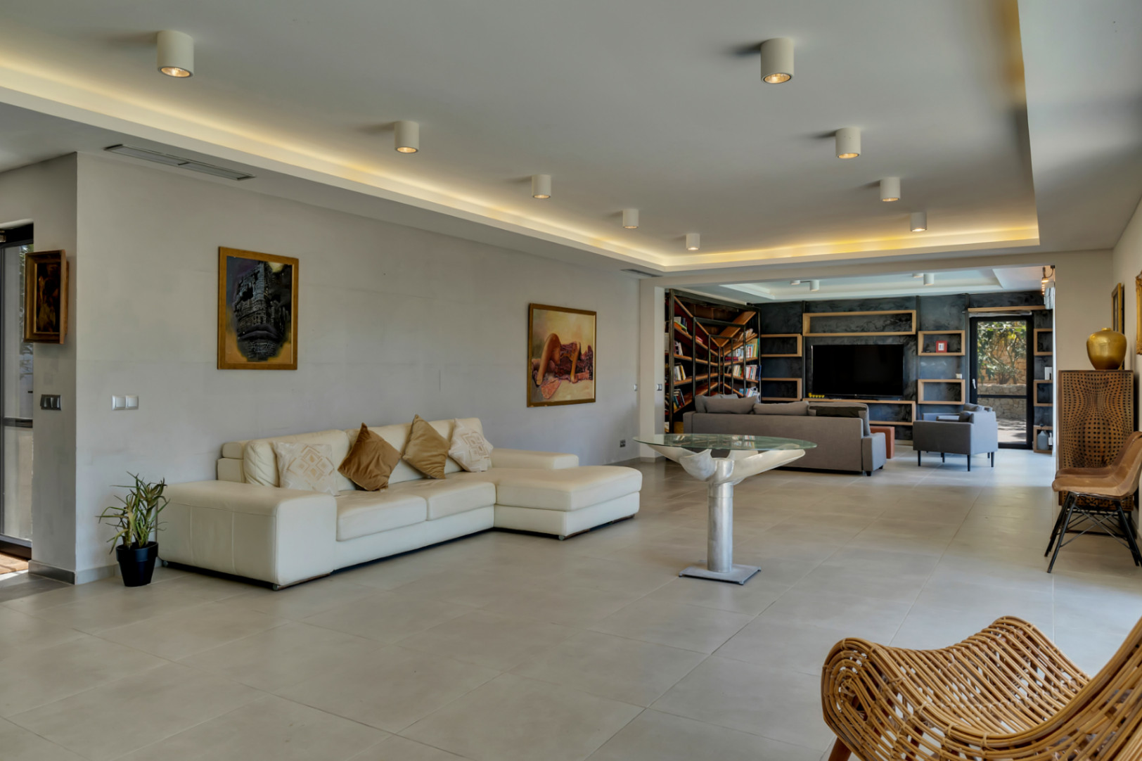 Amplio salón con sofá seccional blanco y mesa de diseño, conectado a una zona de biblioteca y TV con pared oscura y estanterías de madera.