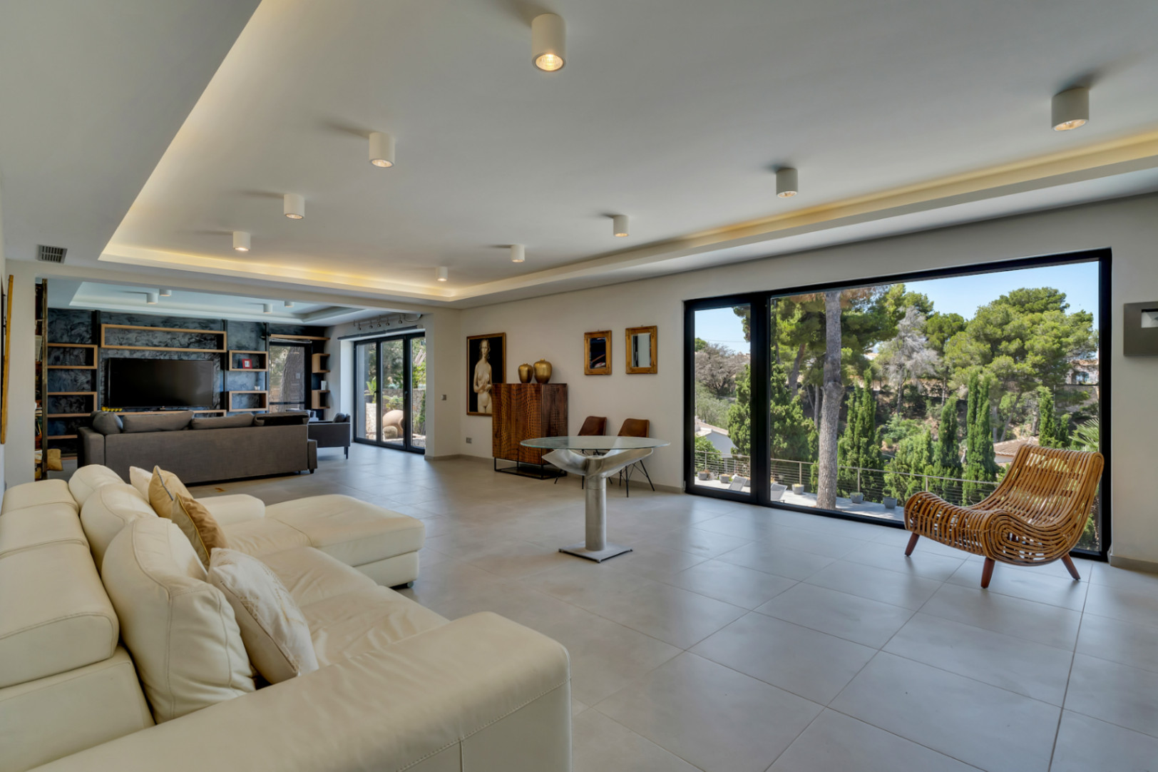 Amplio salón de concepto abierto con grandes ventanales, ofreciendo vistas a la exuberante vegetación y luz natural. Destaca el sofá seccional blanco y la moderna iluminación empotrada.