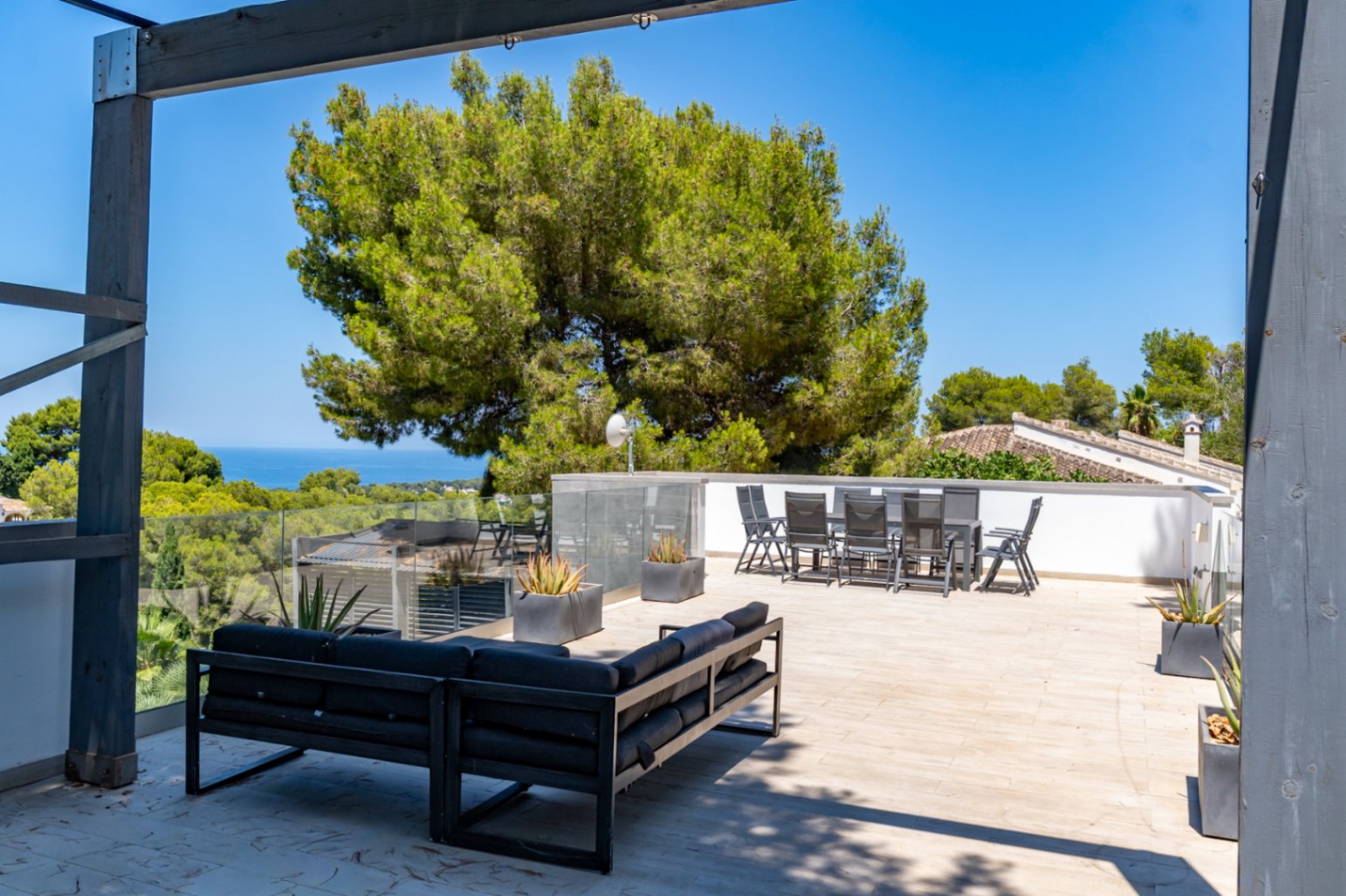 Amplio solárium en la azotea con moderno sofá exterior y conjunto de comedor, barandillas de cristal y vistas panorámicas al mar entre pinos mediterráneos.