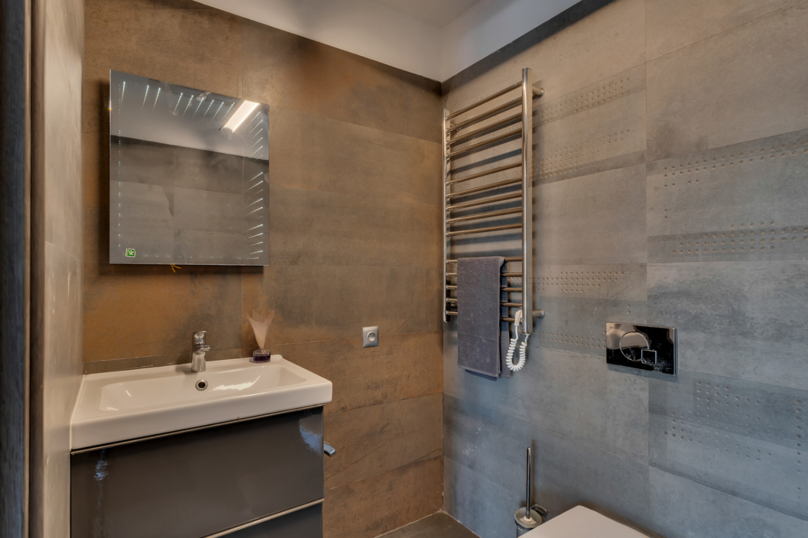Aseo de cortesía con lavabo suspendido sobre mueble gris, espejo iluminado y toallero calefactado. Destacan los azulejos texturizados en tonos grises y óxido.