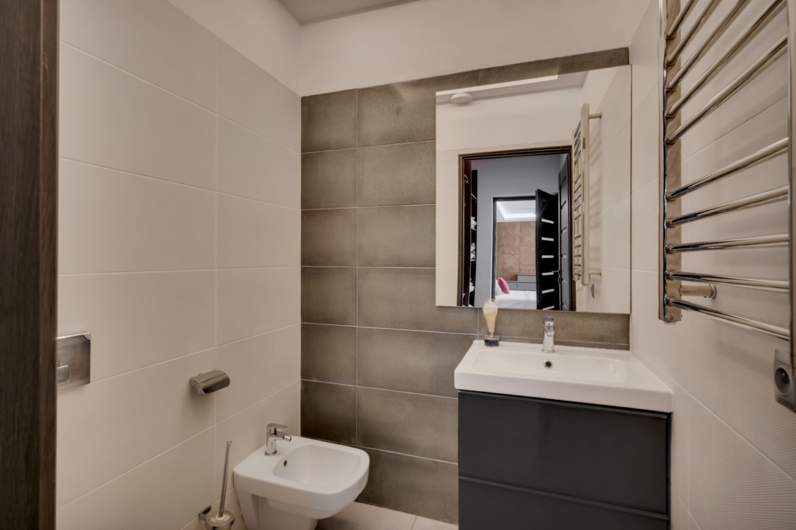 Baño contemporáneo con mueble de lavabo gris oscuro, lavamanos blanco, bidé y toallero calefactado cromado. Las paredes combinan azulejos claros texturizados y oscuros lisos.
