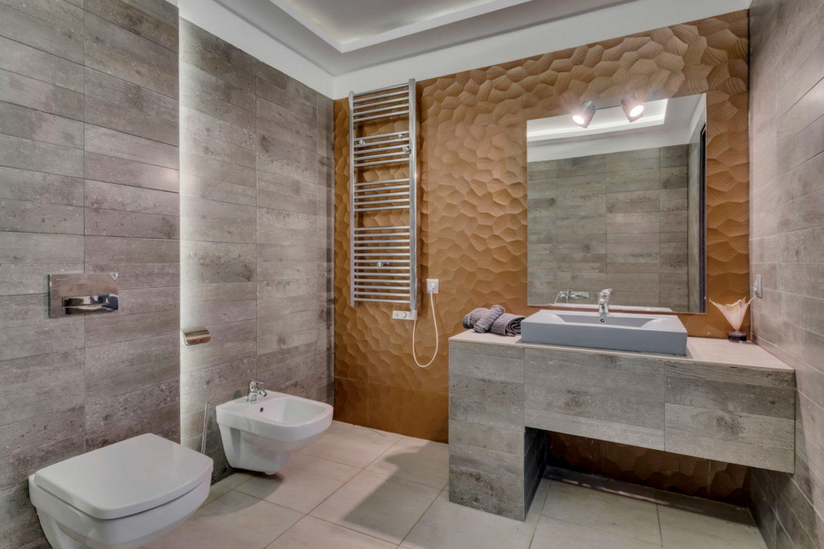Baño moderno con azulejos grises texturizados, una pared decorativa 3D geométrica en tono bronce y un lavabo flotante. Incluye inodoro y bidé suspendidos, además de toallero calefactado.