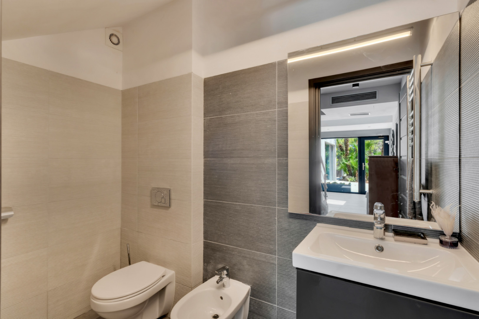 Baño moderno con azulejos texturizados en tonos neutros, inodoro, bidé y lavabo con mueble oscuro. El espejo iluminado refleja un exterior ajardinado.