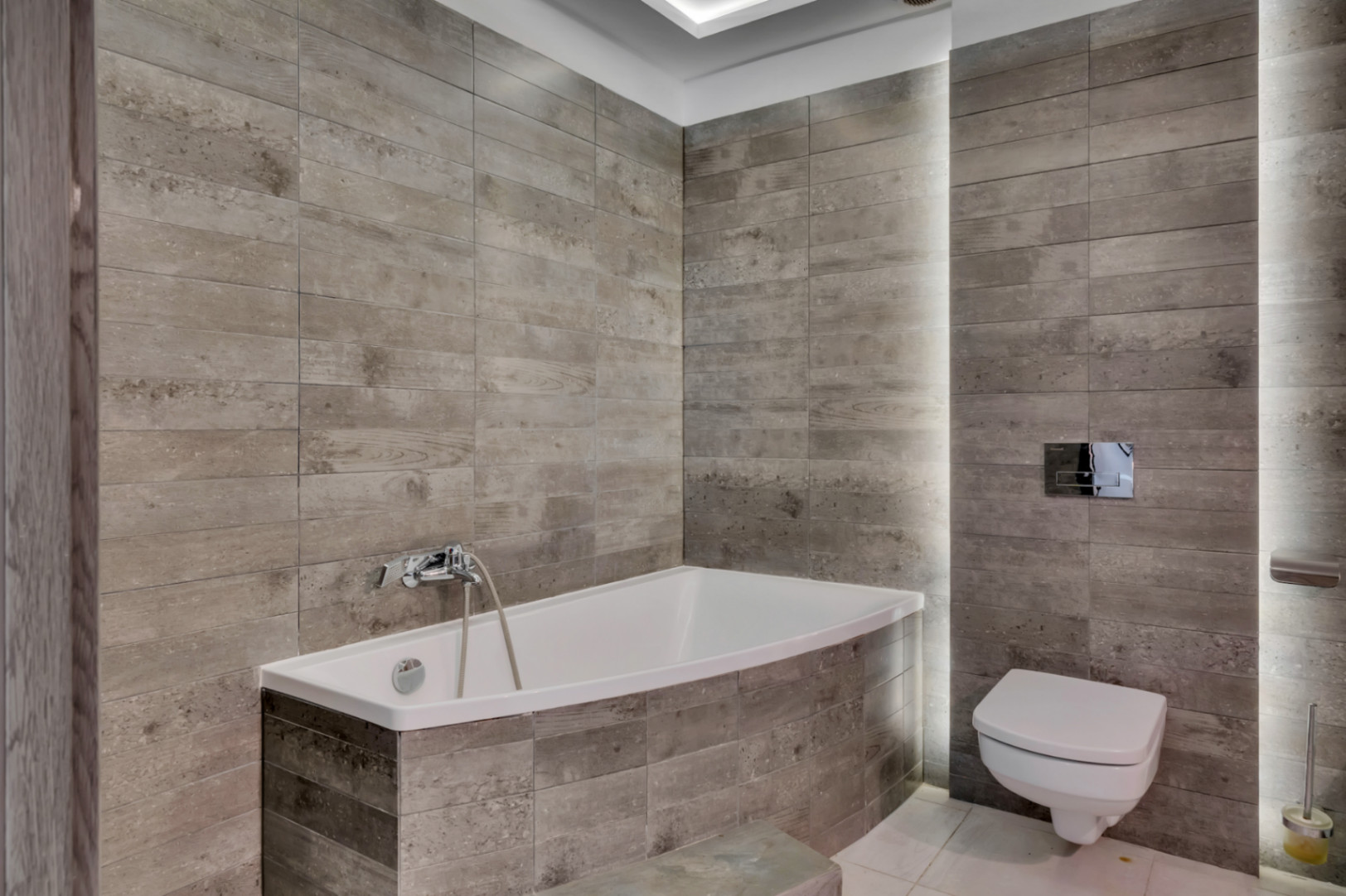 Baño moderno con bañera integrada y WC suspendido, destacando los revestimientos cerámicos grises con textura de madera y la iluminación LED vertical integrada.