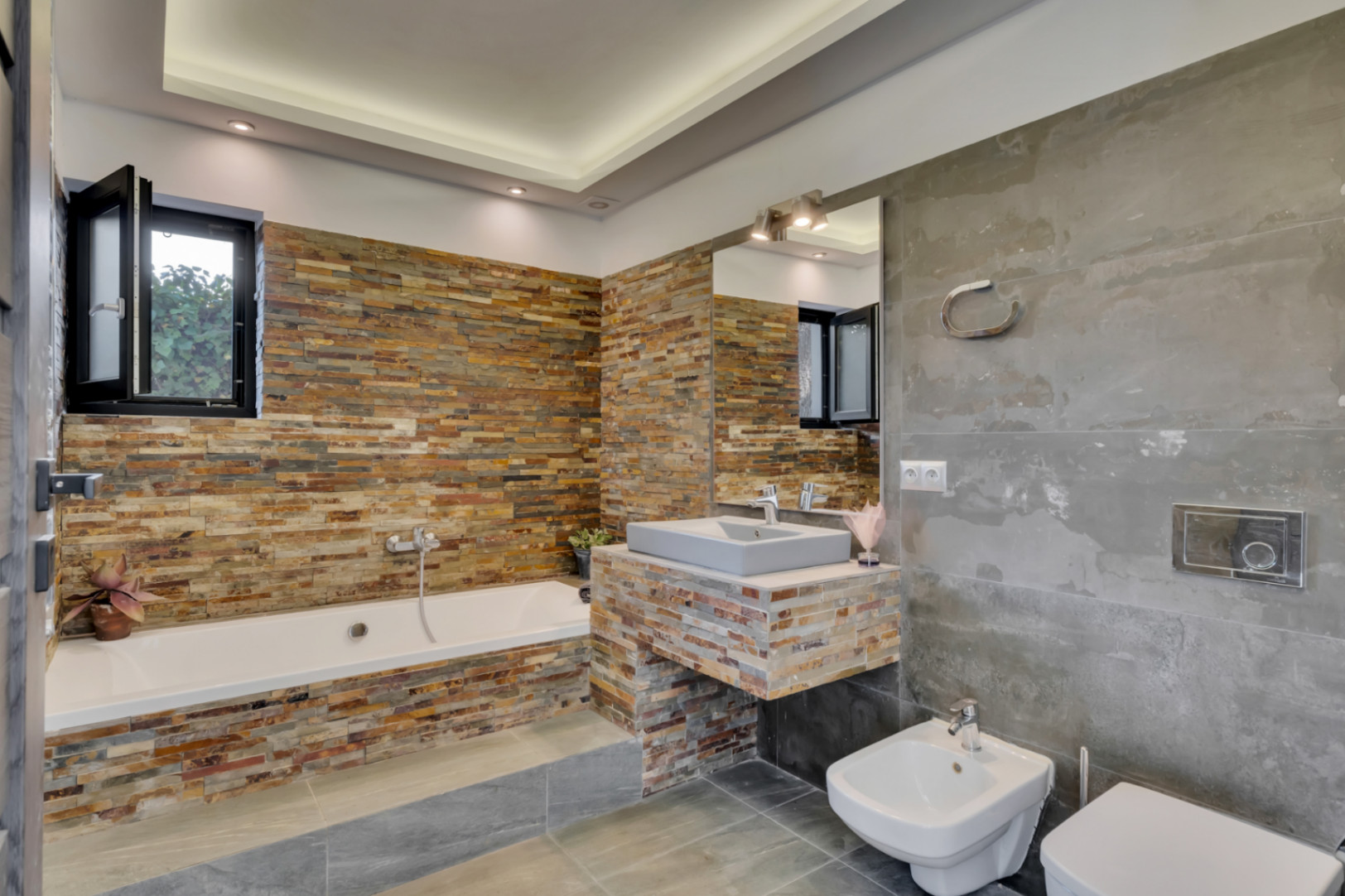 Baño moderno con bañera y lavabo flotante revestidos en piedra apilada multicolor. Grandes azulejos grises cubren paredes y suelo, con iluminación empotrada.