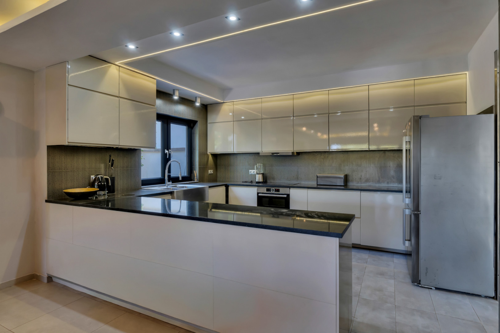 Cocina moderna con armarios de alto brillo color crema, encimeras de granito negro y electrodomésticos integrados. Iluminación LED lineal y empotrada en el techo.