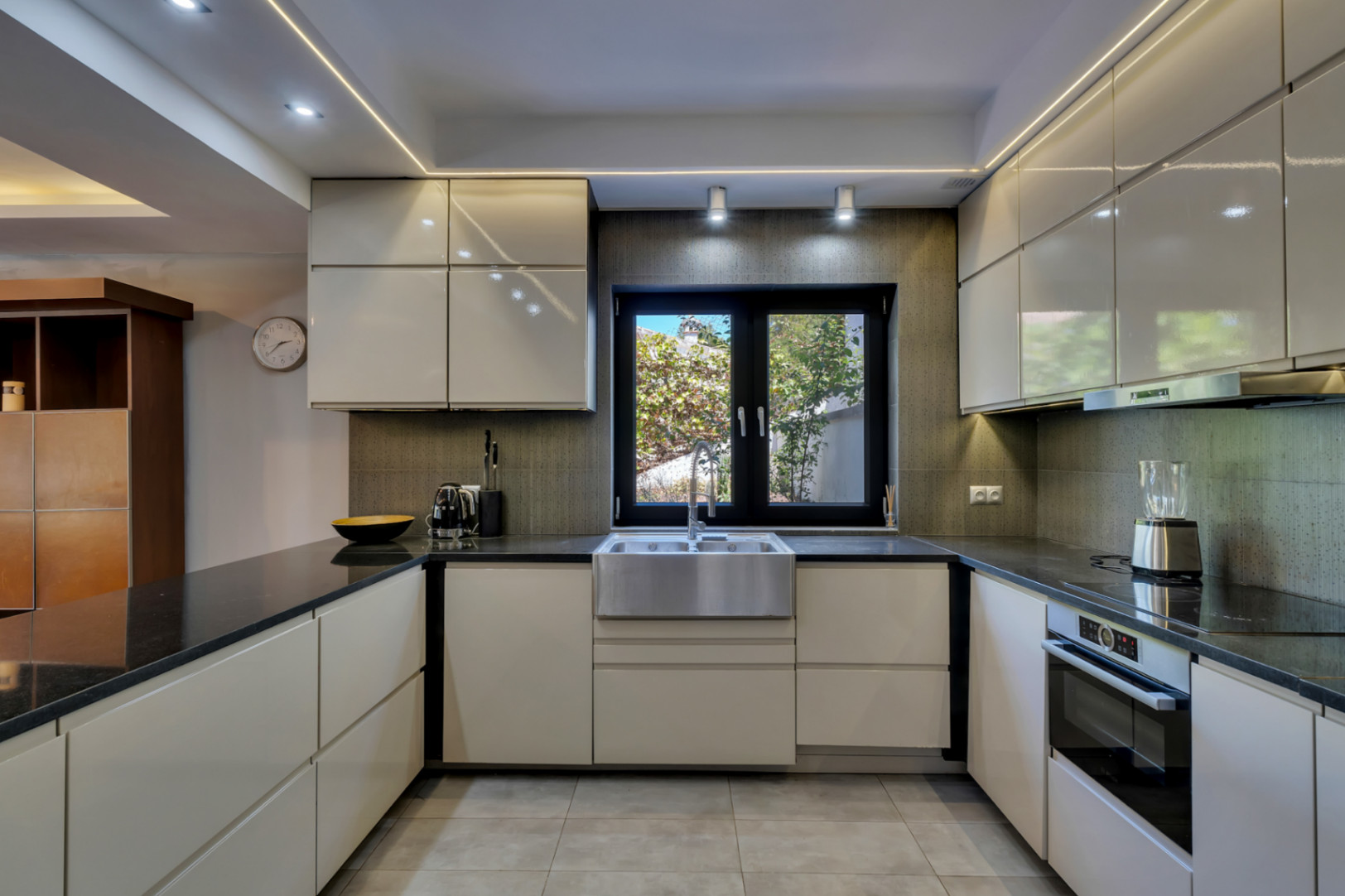 Cocina moderna con armarios lacados en blanco roto, encimeras negras y fregadero de acero inoxidable bajo una ventana que da a la vegetación. Iluminación LED integrada y suelo de baldosas grises.