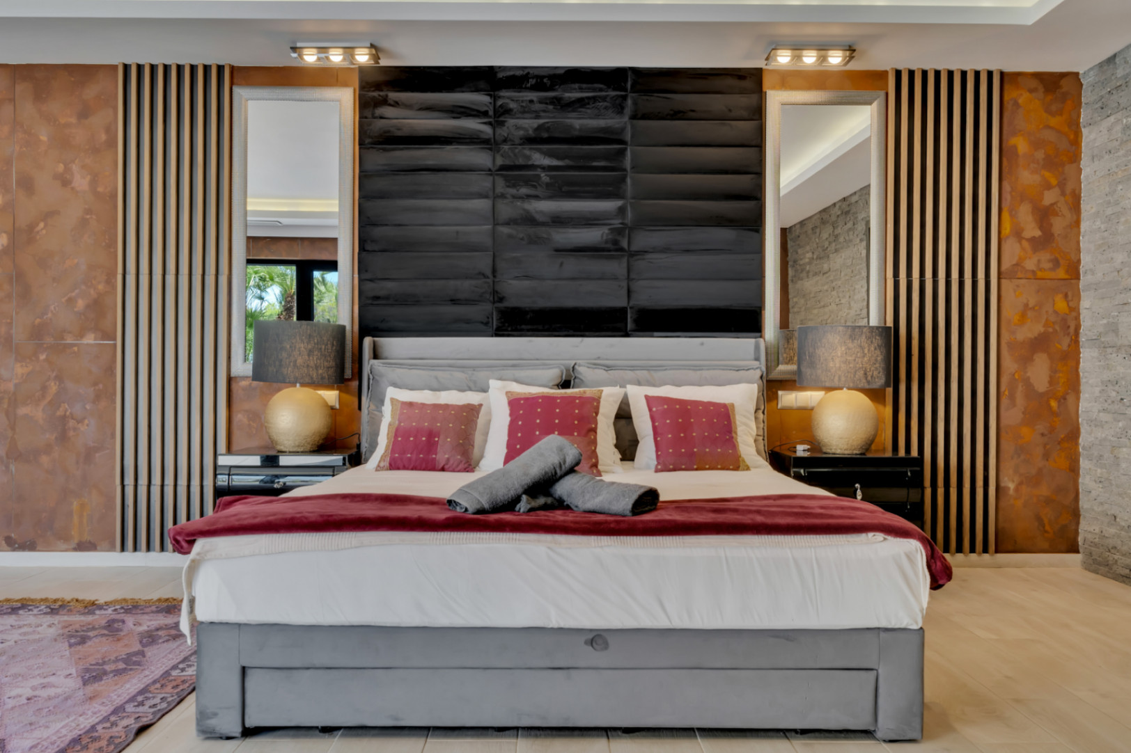 Dormitorio principal con cama tapizada gris, cabecero negro texturizado, espejos y paneles de pared únicos en madera, metal envejecido y piedra. Iluminación a medida crea un ambiente sofisticado.