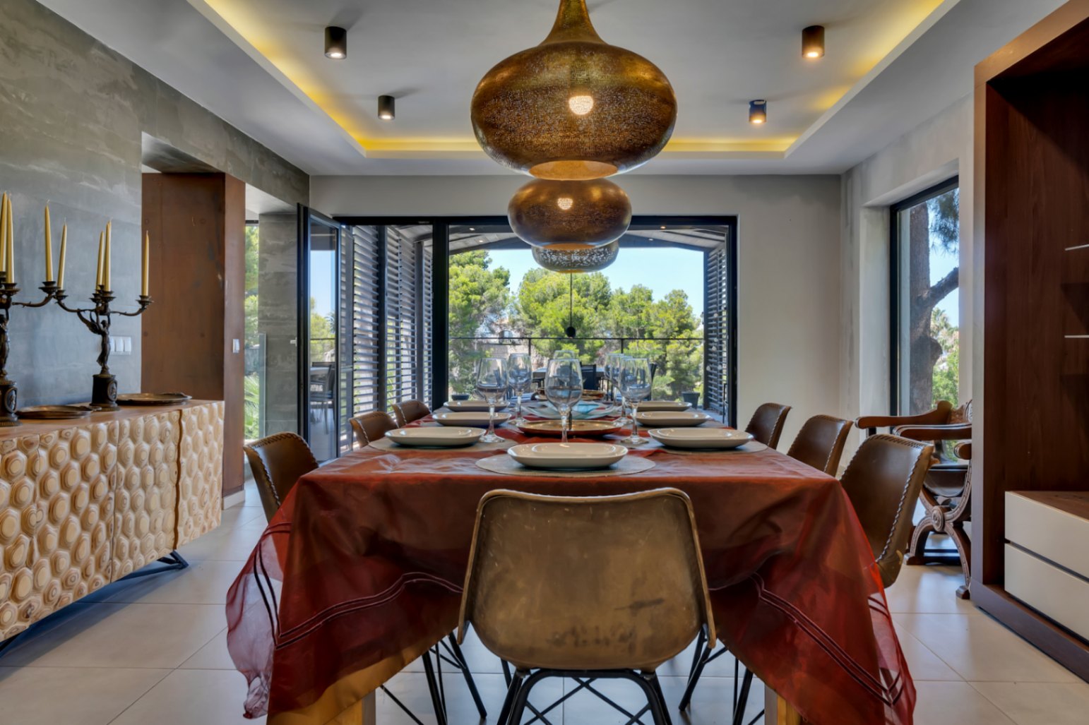 Elegante comedor con lámparas colgantes de bronce ornamentadas, un aparador de madera texturizada y amplios ventanales con vistas serenas a la exuberante vegetación.