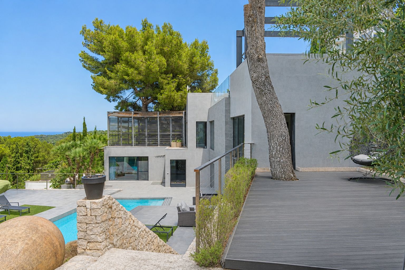 Exterior de villa moderna de varios niveles con fachadas grises, piscina privada y terraza de composite que integra un árbol maduro. Vistas lejanas al mar entre la exuberante vegetación mediterránea.