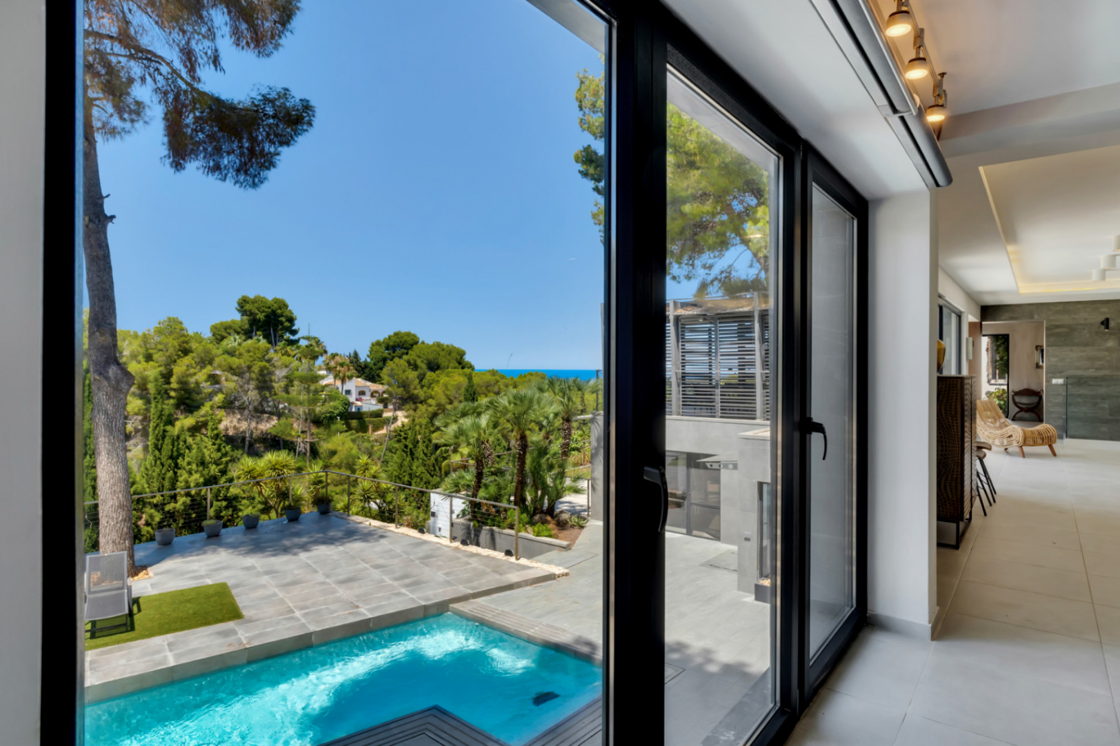 Grandes puertas correderas de cristal negro conectan el interior moderno con la piscina privada, terraza de gres y exuberante paisaje mediterráneo con vistas al mar lejano.