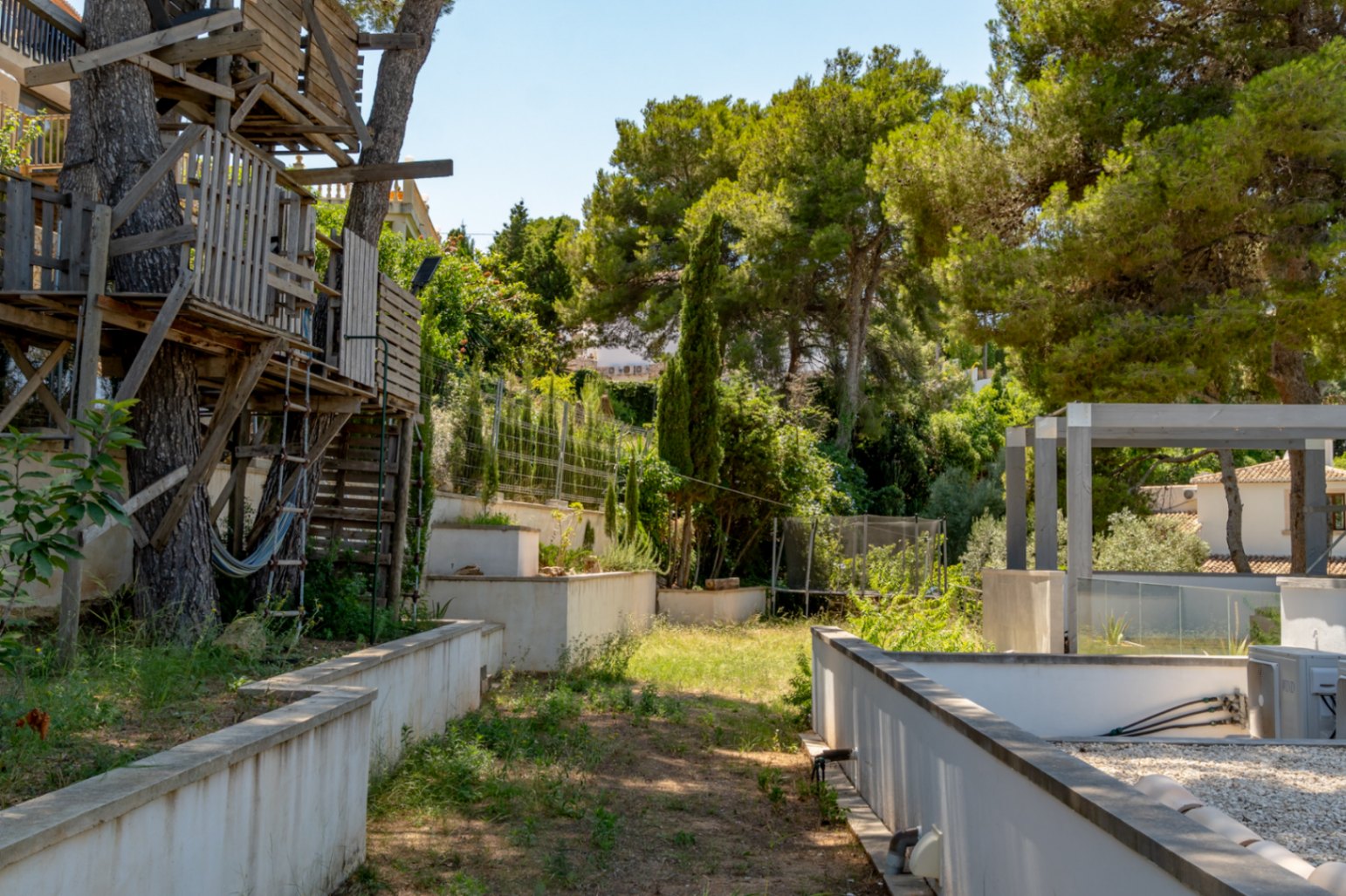 Jardín con una casa en el árbol de madera rústica entre pinos maduros y una hamaca. El espacio incluye muros de contención, vegetación densa y una pérgola moderna.