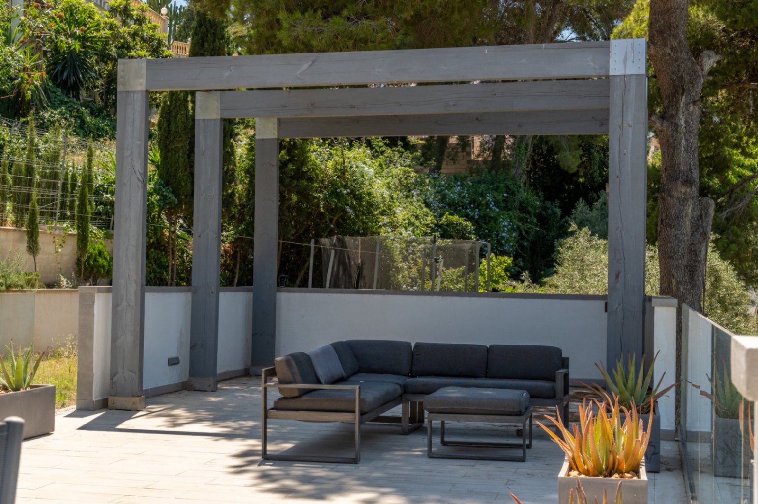 Moderna zona de estar exterior bajo una pérgola de madera gris, con sofá en L y otomana sobre pavimento claro, rodeada de vegetación mediterránea y plantas suculentas en macetas.