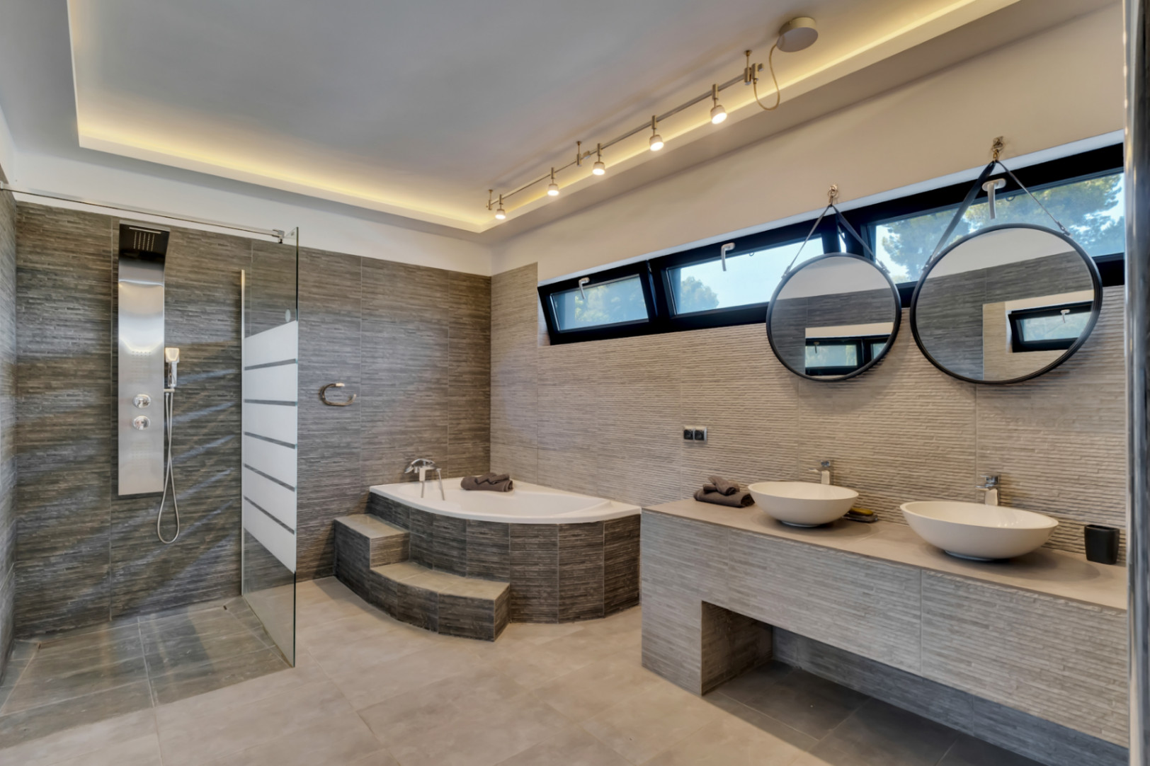 Moderno baño con ducha de hidromasaje, bañera esquinera con escalones y doble lavabo. Azulejos texturizados, espejos redondos y luz natural de ventanas horizontales.