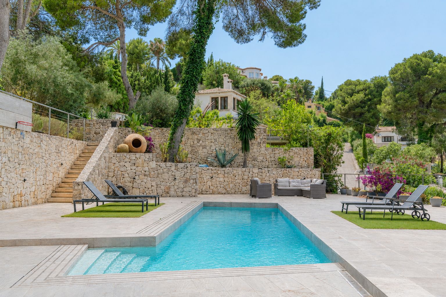 Piscina privada rectangular con escalones integrados, rodeada de pavimento claro y zonas de césped artificial con tumbonas. Muros de piedra natural y vegetación mediterránea en terrazas ajardinadas.