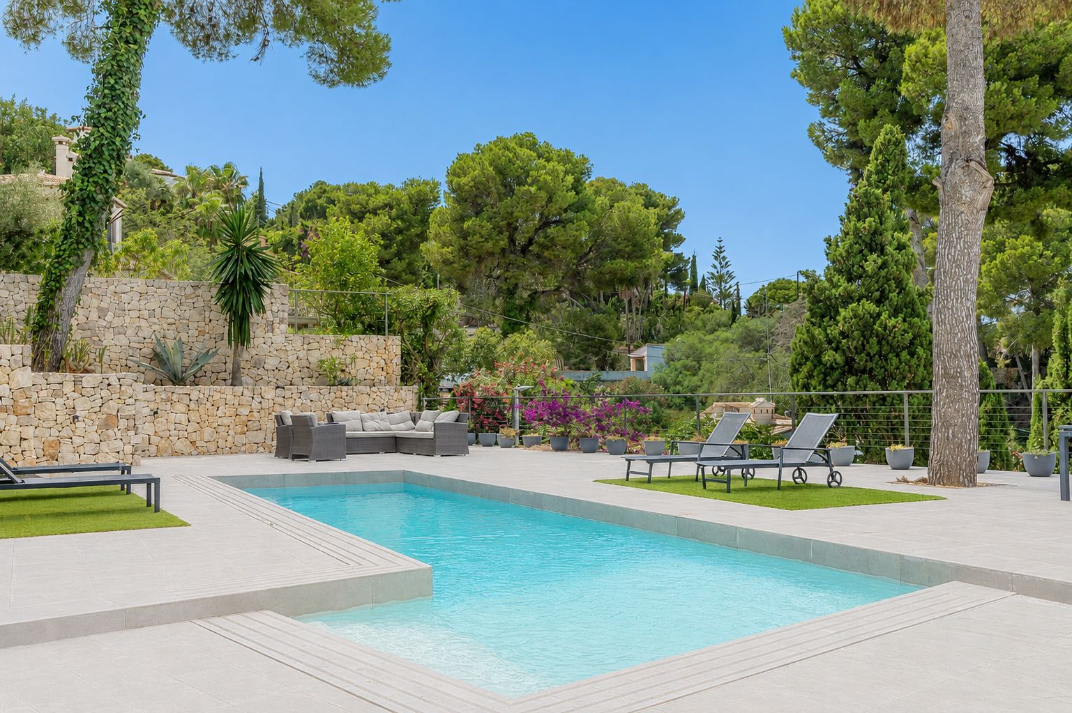 Piscina privada rectangular rodeada de una amplia terraza de gres claro, con zona de relax exterior y muro de piedra natural en un entorno ajardinado.