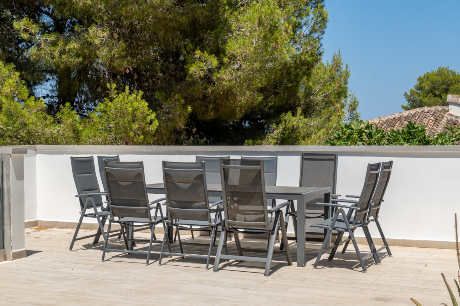 Terraza exterior con mesa de comedor para ocho personas y sillas de diseño moderno, sobre suelo de baldosas claras, enmarcada por una pared blanca y vistas a la vegetación y tejados tradicionales.