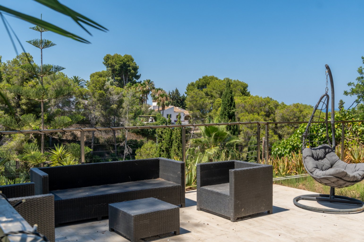 Terraza exterior con mobiliario de ratán oscuro y silla colgante, ofreciendo vistas panorámicas a la exuberante vegetación y una casa tradicional en la distancia.