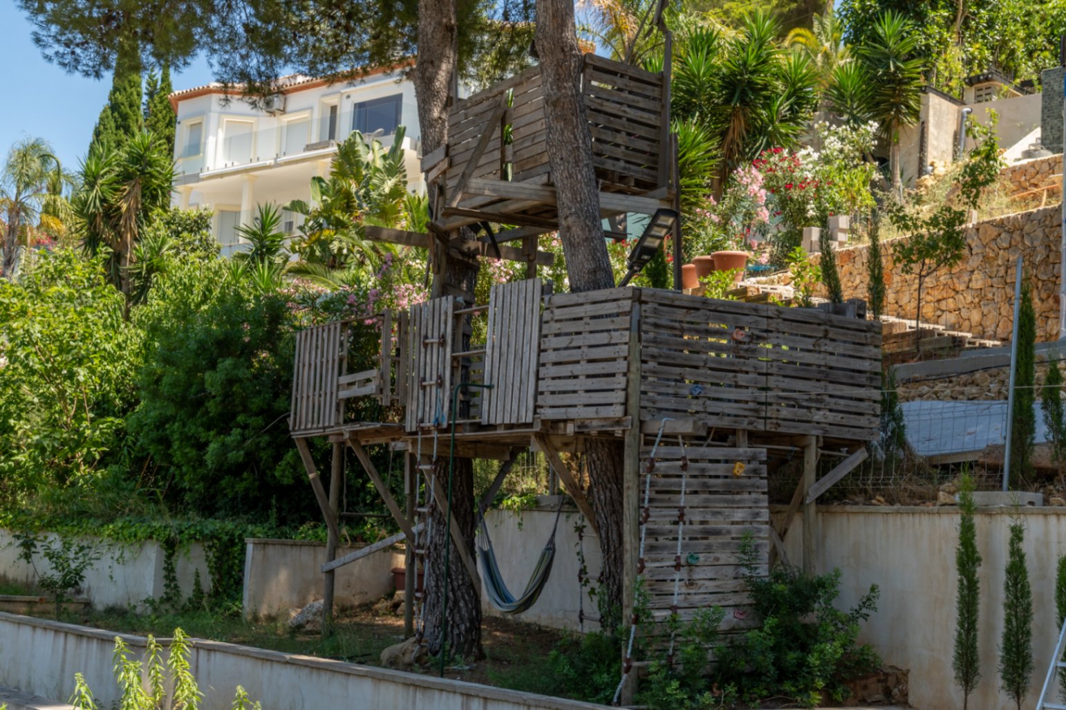 Una rústica casa en el árbol de varios niveles, construida con palets de madera, con escaleras y una hamaca, rodeada de vegetación y una villa moderna al fondo.