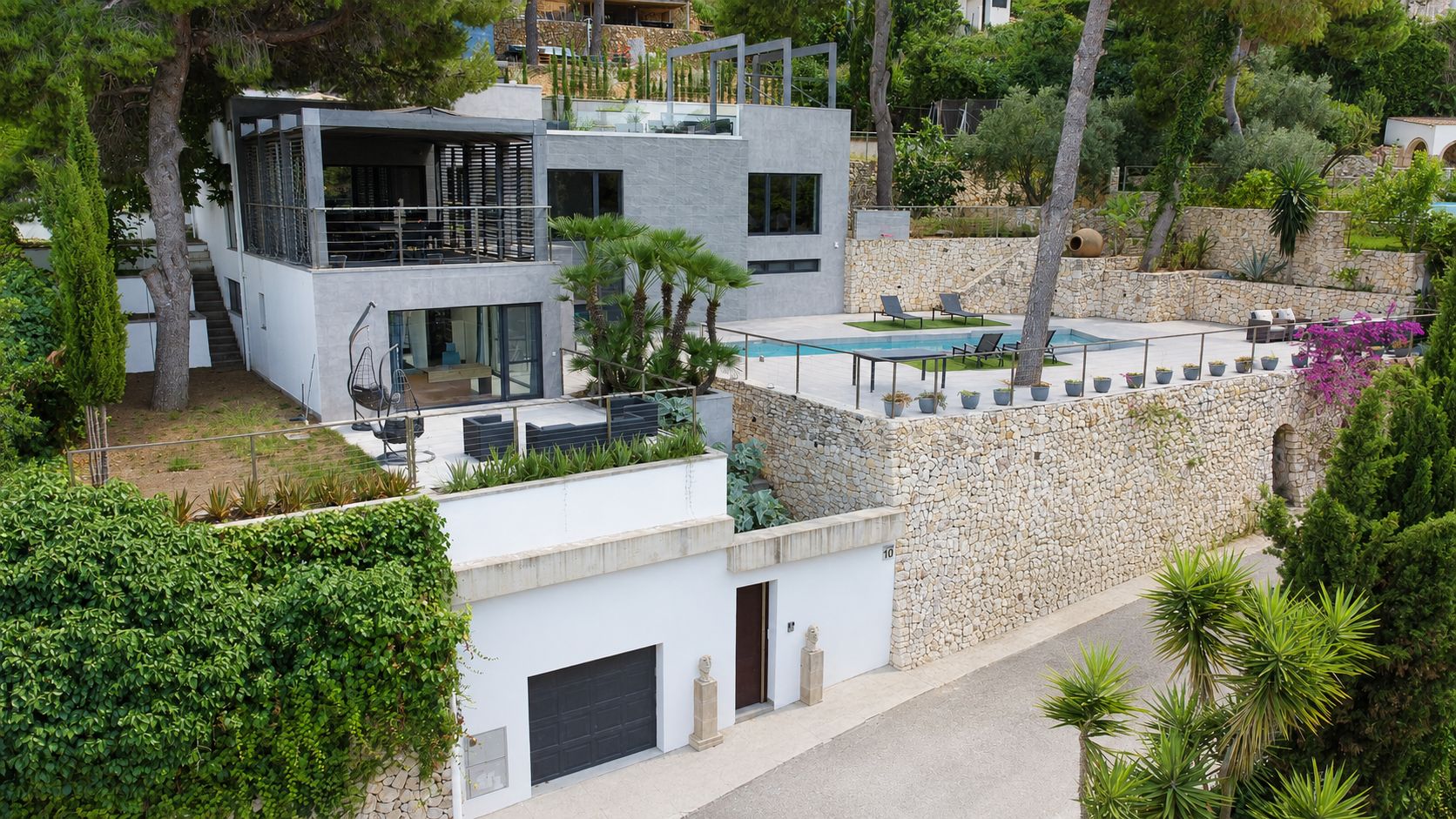 Vista aérea de la villa moderna con piscina privada, terrazas ajardinadas y muros de piedra natural, destacando su diseño multinivel y garaje a pie de calle.