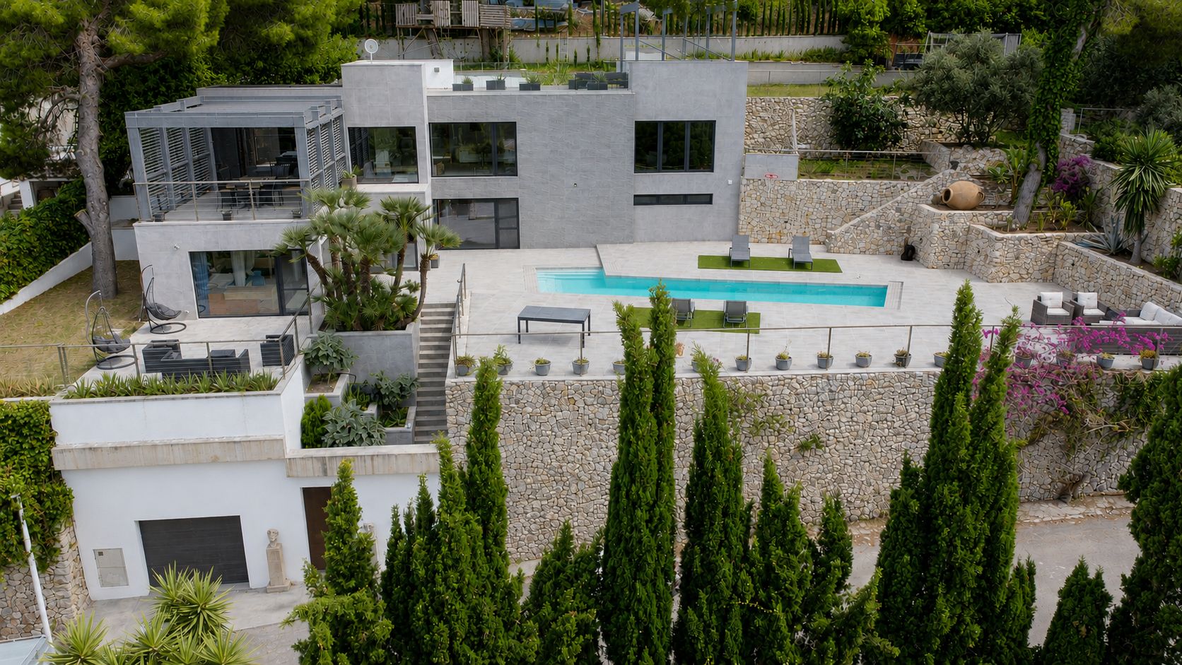 Vista aérea de la villa moderna con piscina rectangular y terrazas escalonadas. Destacan la fachada gris, los jardines en bancales de piedra y las amplias zonas de ocio exterior.