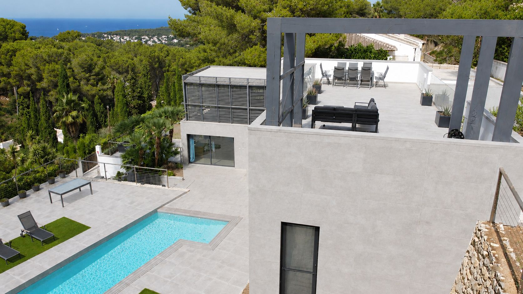 Vista aérea de la villa moderna, destacando la piscina privada de diseño, amplias terrazas de gres y una zona chill-out elevada con vistas al mar Mediterráneo y la vegetación. Diseño contemporáneo.