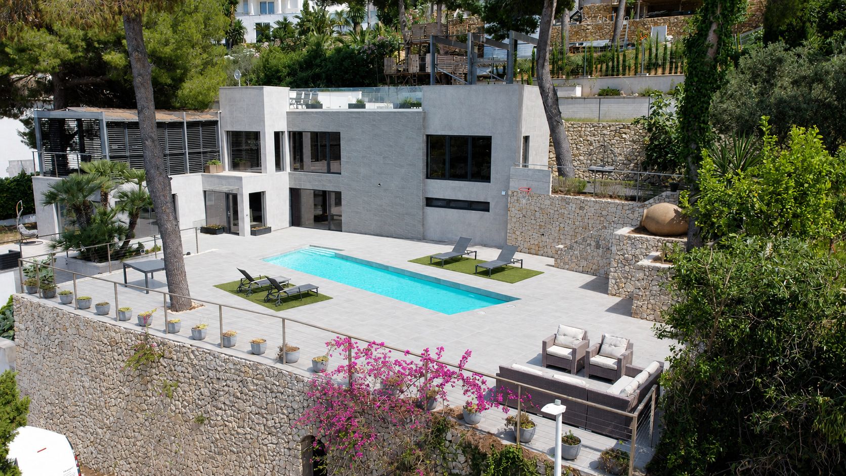 Vista aérea de la villa moderna, destacando su elegante fachada gris, piscina privada rectangular y amplias terrazas de gres. Muros de piedra y vegetación mediterránea enmarcan la propiedad.