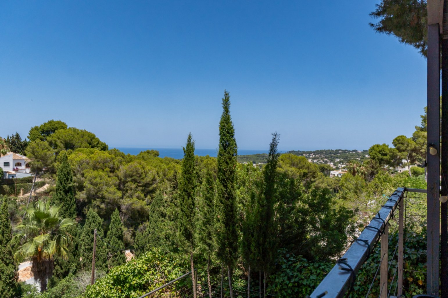 Vista desde la terraza con abundante vegetación mediterránea, cipreses y pinos, el mar a lo lejos y parte de la urbanización de Jávea bajo un cielo azul claro.