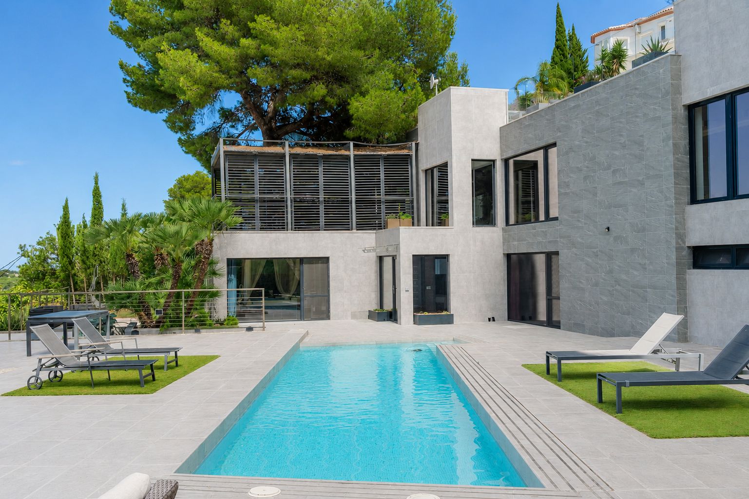 Vista exterior de la villa con piscina privada rectangular, terrazas de gres gris y zonas de césped artificial. Destaca su arquitectura moderna y una terraza superior con lamas.